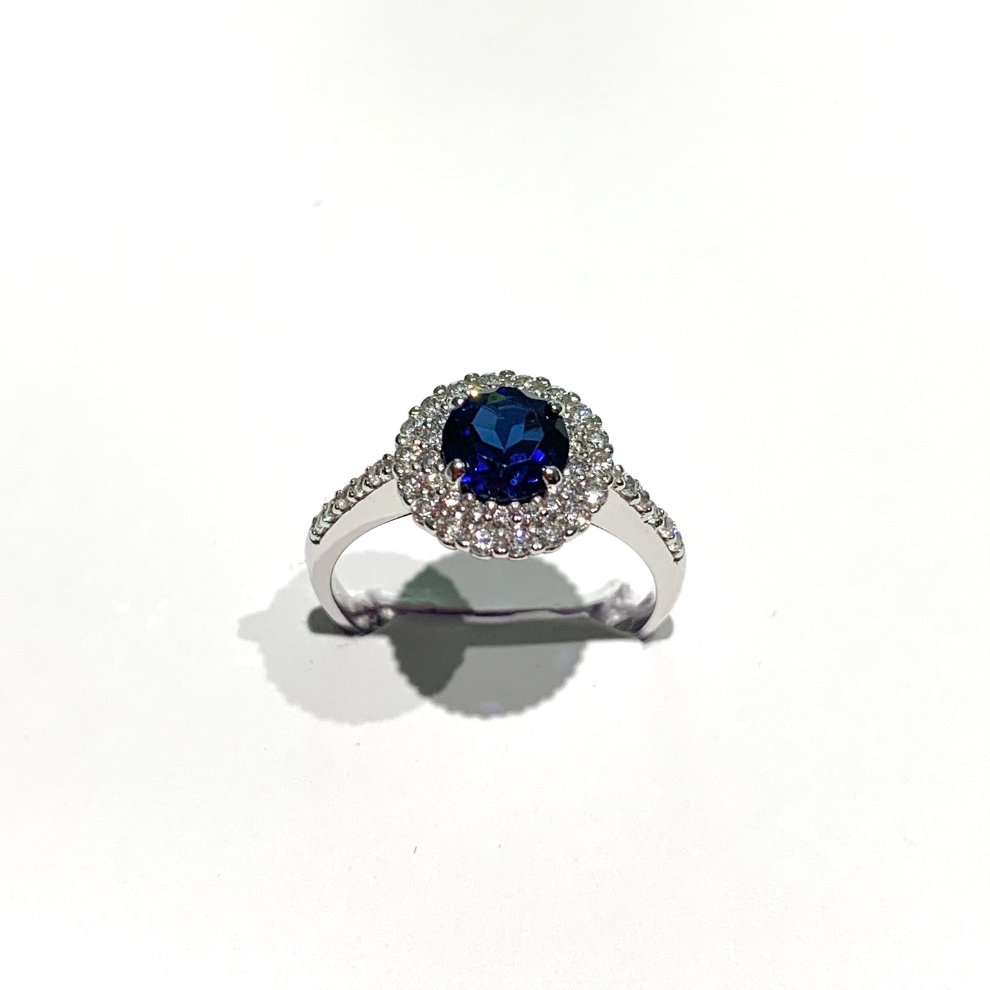 Anello in oro bianco 18kt con zircone centrale tondo colore zaffiro circondato da piccoli zirconi bianchi. Dimensione totale 1 cm. Misura 14.
