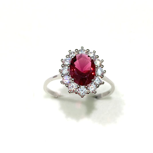 Anello in oro bianco 9kt con zircone colore rubino, circondato da zirconi bianchi. Dimensione zircone 0,5x0,8 cm. Dimensione totale 1 cm. Misura 12.